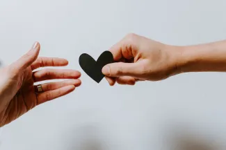 Image of two hands against a white background. One of the hands is passing a black paper heart to the other one