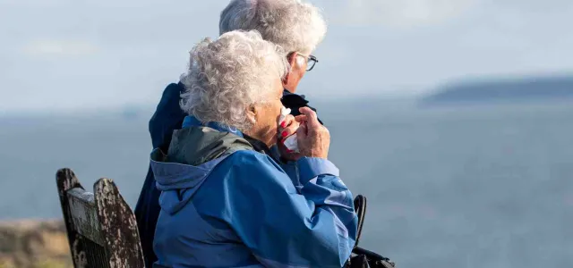 Two elderly people sitting on a bench looking at the sea